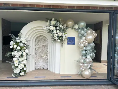 Corporate decor in Loughborough by Lulu Belle Events. Organic balloon garland running from the top of the arch down the right side, interspersed with small white florals and eucalyptus foliage. Chrome silver, sage green, champagne pearl, gold. Organic clustered garland with mixed balloon sizes including oversized chrome and standard balloons.
