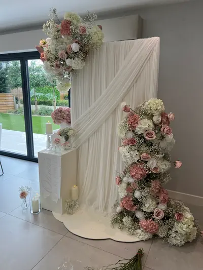 Baby shower decor in Barrow upon Soar by Lulu Belle Events. White, dusky pink, and blush with greenery accents. Floral cluster at base of backdrop continuing the cascade, comprising dusky pink roses, white hydrangeas, pink hydrangeas, and gypsophila. Indoor private venue with large sliding glass doors overlooking a garden patio.