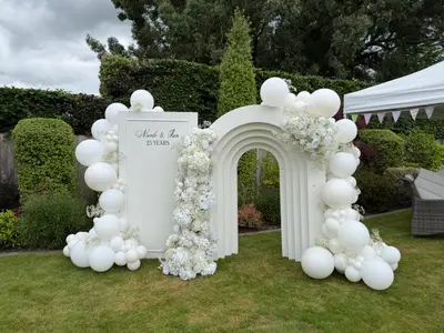 Event styling & planning in Kirby Muxloe by Lulu Belle Events. Outdoor garden with manicured lawn, hedging and topiary. All white. Organic balloon garland in all white with mixed sizes from large round orbs to small clusters, wrapping around both the flat backdrop and the arch structure.