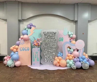 Birthday decor in Cosby by Lulu Belle Events. Pastel pink, lilac, peach, mint green, baby blue, silver accents. Retro groovy / Y2K style with smiley face, peace sign, lightning bolt and daisy motifs. Indoor hall with parquet wood flooring, grey painted walls and archway recess.