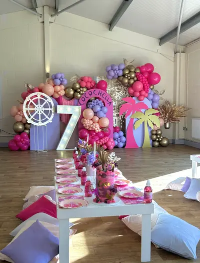 Birthday decor in Leicester by Lulu Belle Events. Coachella-inspired festival theme with tropical and disco elements. Hot pink, lilac/lavender, peach, gold, white. Indoor hall with white painted breeze-block walls, apex roof with exposed steel beams, and wooden parquet flooring. White Ferris wheel prop/frame positioned to the left of the backdrop.