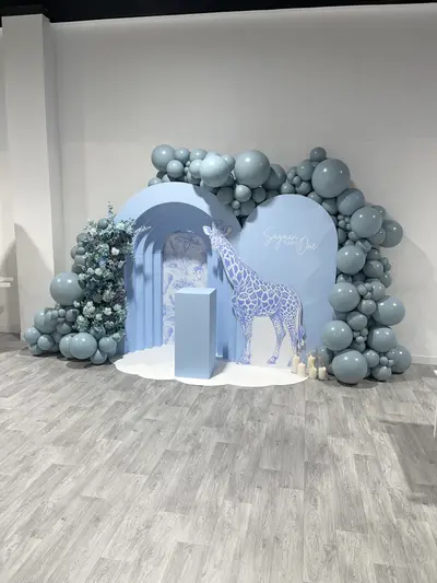 First birthday decor in Leicester by Lulu Belle Events. Dusty blue, slate blue, white, with muted blue-toned florals. Safari and giraffe with blue toile aesthetic. Large organic balloon garland in dusty blue and slate tones wrapping around the full width of the backdrop structure, from floor level on both sides up and over the top. Dusty blue, slate blue, muted teal, varied sizes.