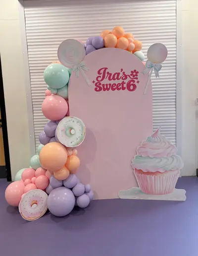 Birthday decor in Leicester by Lulu Belle Events. Sweet shop / candy / pastel desserts. Pastel pink, lilac, peach, mint green. Arch-top panel, flat-faced freestanding structure. Single organic balloon garland running up the left side and across the top of the arch panel.
