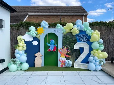 Birthday decor in Narborough by Lulu Belle Events. In the Night Garden. Outdoor residential garden patio. Navy blue, sky blue, mint green, yellow-green, and white. Igglepiggle plush toy seated on a swing inside the arched centre panel of the backdrop.