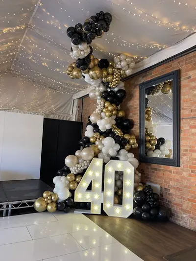 Birthday decor in Southwell by Lulu Belle Events. Black, chrome gold, and ivory/cream. Large organic balloon garland column rising from floor to ceiling, incorporating the light-up numbers at the base, with balloons spilling out at ground level on both sides. Organic, dense cluster style, floor-to-ceiling column. Black, chrome gold, ivory/cream, and matte white.