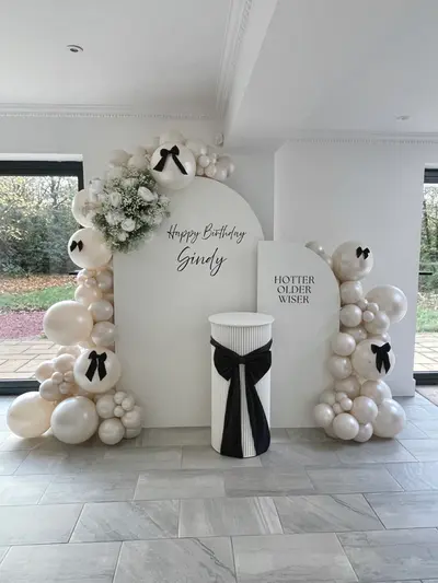 Birthday decor in Lutterworth by Lulu Belle Events. Ivory, cream, white and black. Organic balloon garland running along the top of the backdrop arch and down both sides. Ivory, cream, and pearl white in mixed sizes. Indoor event space with white walls, cornice detailing, and large windows overlooking a garden.