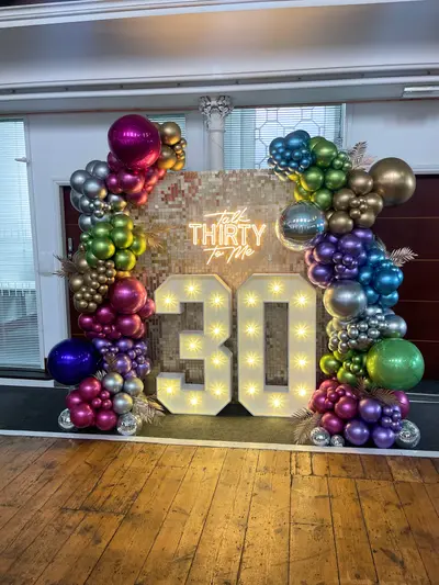 Birthday decor in Burton upon Trent by Lulu Belle Events. Indoor function room or hall with white ceiling, large windows and a column visible. Two large organic balloon garland columns framing either side of the backdrop, extending across the top.