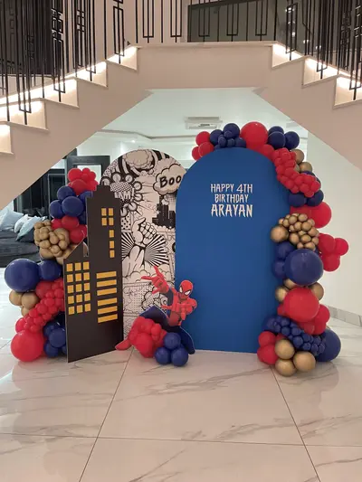 Birthday decor in Oadby by Lulu Belle Events. Spider-Man / comic book superhero. Organic balloon garland framing the arched blue panel and extending down both sides and across the top. Navy blue, red, and gold/bronze. Mixed-size organic clusters forming a full arch around the blue panel with ground-level clusters at the base.