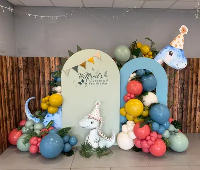 Christening decor in Ashby-de-la-Zouch by Lulu Belle Events. Theme Dinosaur. Large organic balloon garland wrapping around and between both arch panels and pooling at ground level on both sides. Dusty blue, sage green, coral/pink, mustard yellow, cream/white. Sage green, dusty blue, mustard yellow, coral pink, cream and white.