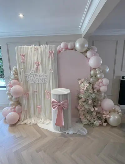 First birthday decor in Rothley by Lulu Belle Events. Soft pink, ivory/cream, white and champagne/pearl. Organic balloon garland wrapping around the arched panel and extending to the floor on both sides. Pale pink, pearl/ivory and white in mixed sizes including oversized white/cream orbs.