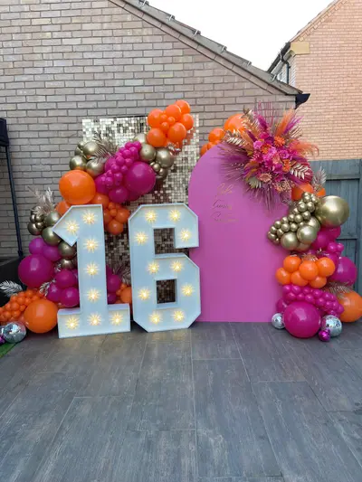 Birthday decor in Broughton Astley by Lulu Belle Events. Outdoor residential garden or patio area. Hot pink, orange, chrome gold, silver. Large organic balloon garland wrapping around both sides of the backdrop display, incorporating the light-up numbers. Organic cluster style with varied balloon sizes from small to large.
