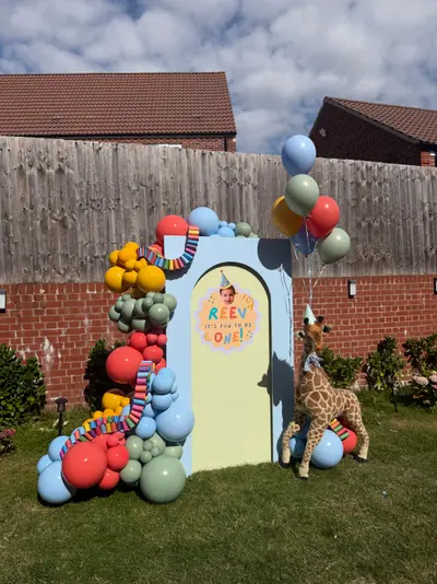 First birthday decor in Leicester Forest East and Kirby Muxloe by Lulu Belle Events. Large organic balloon garland wrapping up the left side and across the top of the arch structure. Baby blue, red/coral, mustard yellow, sage green, and mixed multicolour small accent balloons.