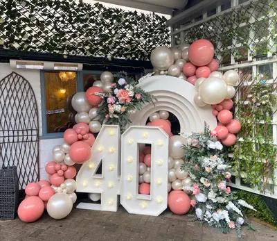 Birthday decor in Kibworth Beauchamp by Lulu Belle Events. Large organic balloon garland wrapping around the arch structure and light-up numbers. Dusty rose/terracotta pink, champagne/cream, and pearl white in mixed sizes including oversized orbs. Organic garland style with varied balloon sizes from small clusters to large statement orbs.