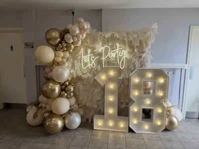 Event styling & planning in Leicester by Lulu Belle Events. Indoor function room with tiled floor and painted grey/white walls. White, cream, and gold (including chrome gold). Organic balloon garland in cream, white, and chrome gold balloons of mixed sizes, positioned to the left side of the backdrop and light-up numbers.