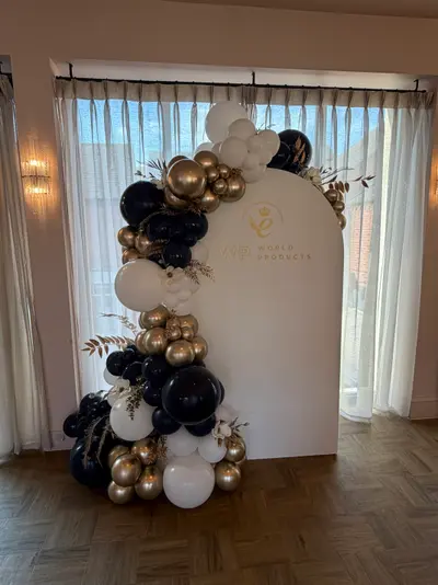 Corporate decor in Leicester by Lulu Belle Events. Arch-top panel with rounded arch at top and flat base. Navy, chrome gold, and white. Single organic balloon garland running from base up the left side and across the top of the arch backdrop. Navy/black, chrome gold, and white.