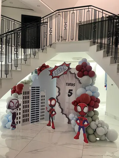 Birthday decor in Oadby by Lulu Belle Events. Spidey and His Amazing Friends (Spiderman). Rectangular panel with shaped cut-out extensions depicting city buildings on the left side. Large organic balloon garland arching over and around the backdrop. Dark red/burgundy, light blue/pale blue, sage green/grey-green, and white.