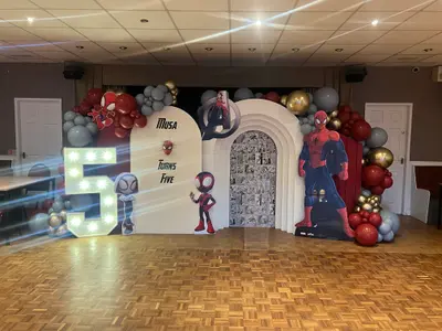 Birthday decor in Rugby by Lulu Belle Events. Spider-Man and Marvel superheroes. Indoor function room or social club with suspended tile ceiling, recessed spotlights, and parquet wood floor. Organic balloon garland running along the top of the full backdrop width and down both sides. Red, grey/slate, chrome gold, and chrome silver in mixed sizes.