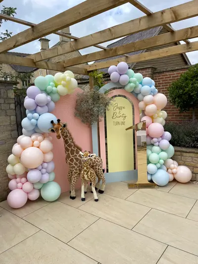 First birthday decor in Melton Mowbray by Lulu Belle Events. Full organic balloon garland arch framing the entire backdrop. Lilac, mint green, baby blue, peach, blush pink, lemon yellow, white. Organic clustered garland with mixed balloon sizes from small to large. Two large plush giraffe figures, one adult-sized and one smaller calf-sized, placed in front of the backdrop.