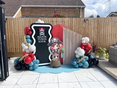 Birthday decor in Narborough by Lulu Belle Events. Theme Pirate. Small barrel prop at base of character cutout, draped with fishing net. Outdoor residential garden patio. Organic balloon garland flanking both sides of the backdrop display.