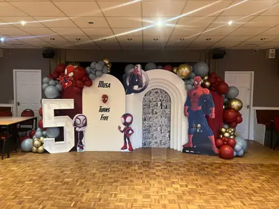 Birthday decor in Shilton and Barnacle by Lulu Belle Events. Spider-Man / Marvel. Red, grey, white, gold, black. Indoor function room or community hall with suspended tile ceiling and parquet wood floor. Organic balloon garland running across the top and sides of the backdrop and around the light-up number.