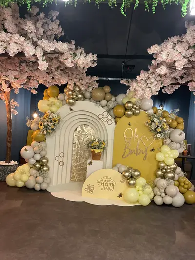 Baby shower decor in Leicester by Lulu Belle Events. Honey pot prop labelled 'HUNNY' used as a floral vessel inside the arch, filled with sunflowers and white blooms. Pink cherry blossom artificial trees framing the backdrop on the left side and overhead, with trailing green foliage hanging from the ceiling.