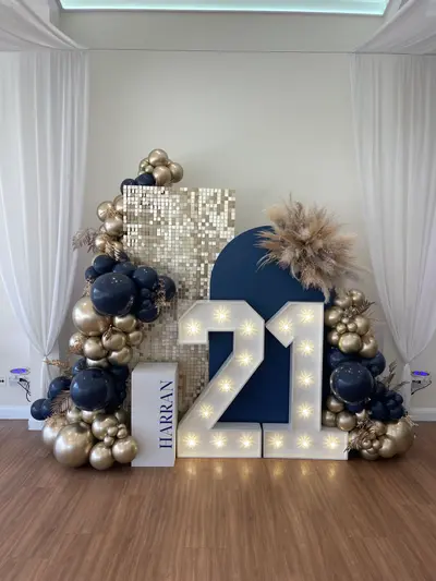Birthday decor in Leicester by Lulu Belle Events. Navy blue, gold chrome, and white. Organic balloon garland arching across the full display, flanking both sides. Navy blue matte and gold chrome in mixed sizes. Indoor function room with white walls and a skylight ceiling.