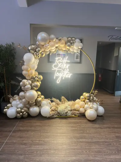 Wedding decor in Syston by Lulu Belle Events. Organic balloon garland wrapped around the circular hoop arch, concentrated on the left side and top, trailing to the base on both sides. White, ivory/cream, gold chrome, and silver chrome. Organic cluster style with mixed balloon sizes including large statement balloons and small filler balloons.