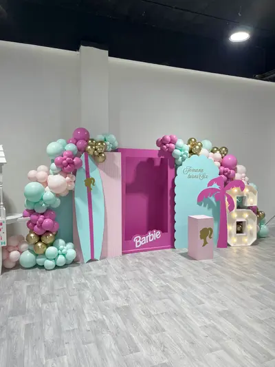 Birthday decor in Leicester by Lulu Belle Events. Event theme Barbie. Hot pink, mint green, baby pink, and gold chrome. Organic cluster style forming an arch across the full width of the backdrop display. Teal and pink striped surfboard cutout with gold Barbie silhouette, positioned to the left of the Barbie box backdrop.
